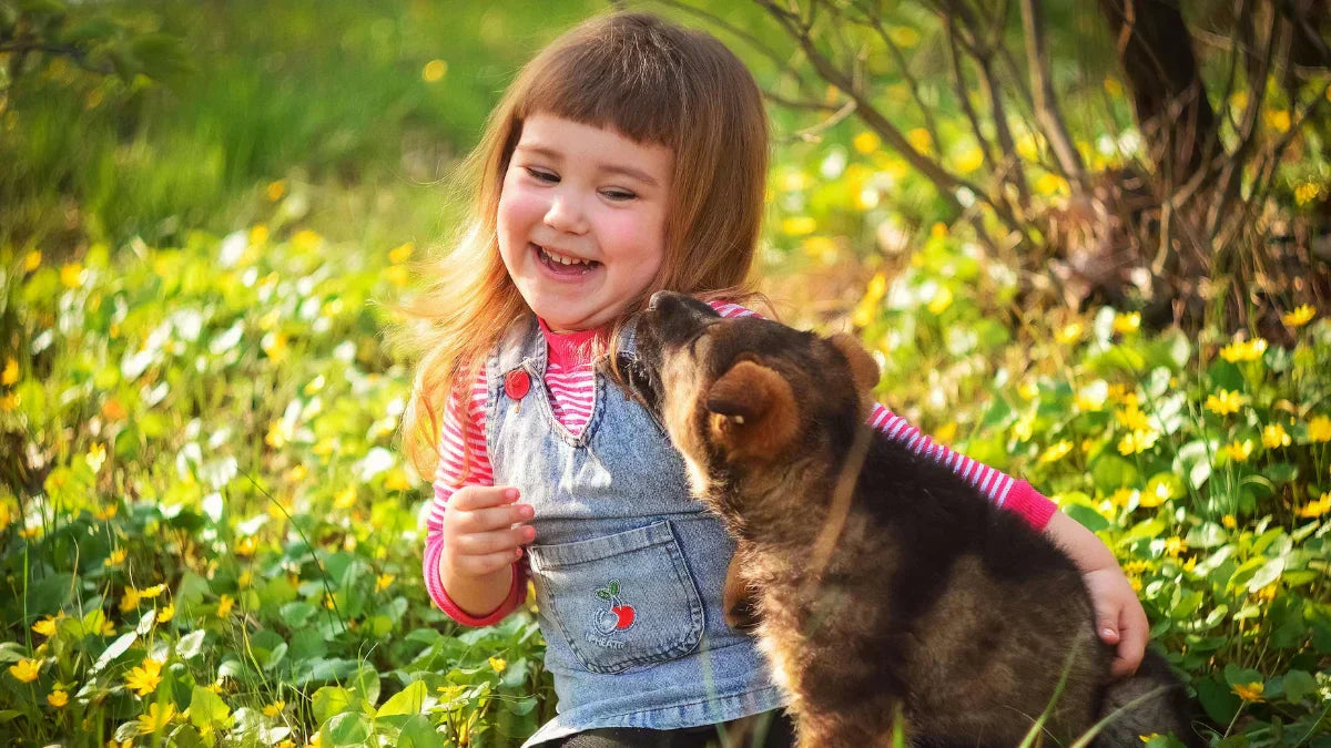 Niña riendo mientras un perro la lame en el rostro, en un jardín al aire libre durante el día.