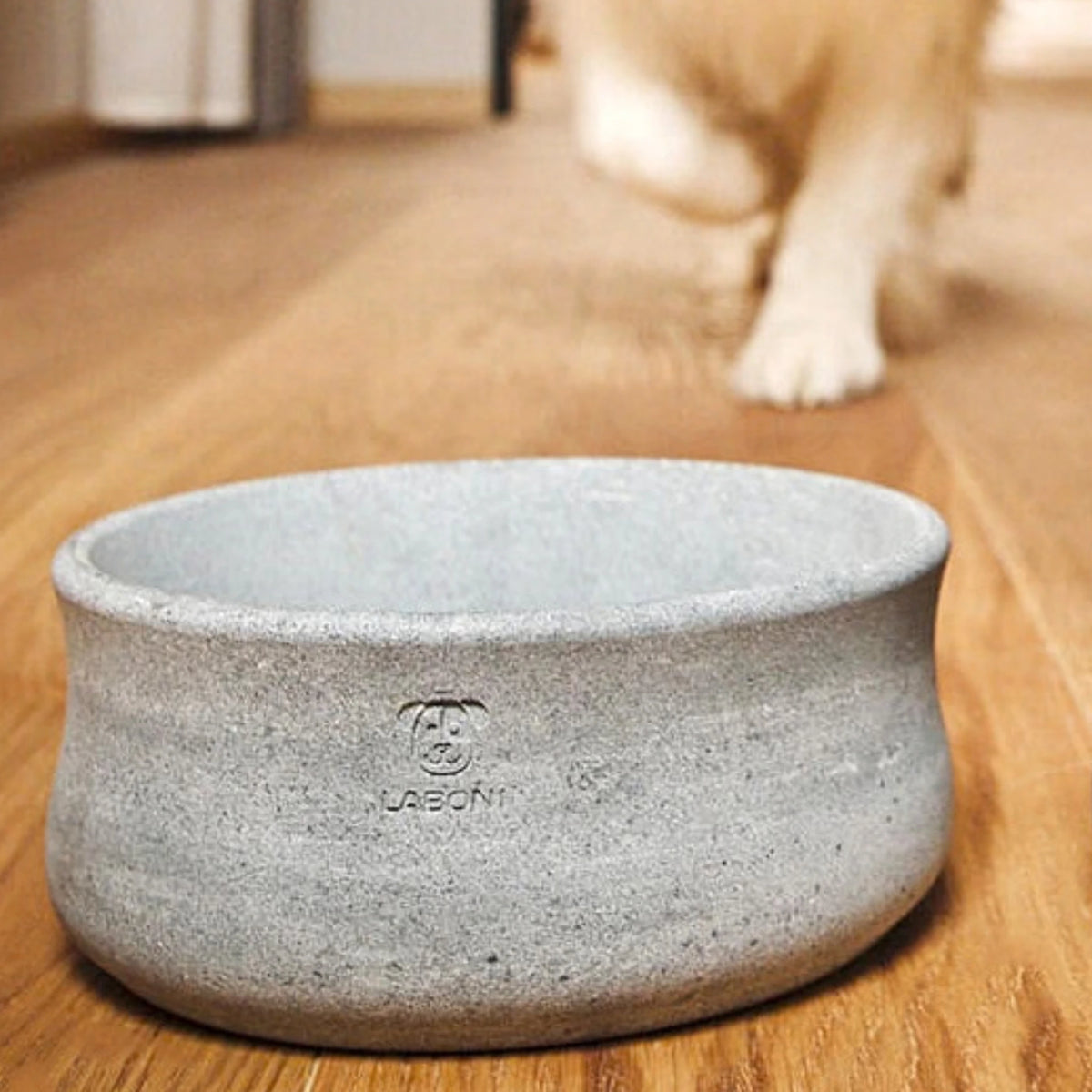 Comedero y bebedero ROCKY de LABONI en piedra natural gris, sobre suelo de madera con un perro al fondo.