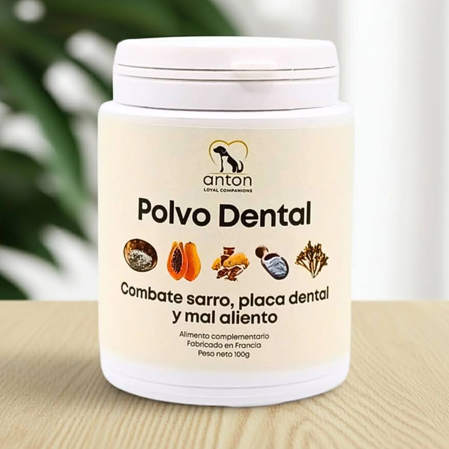 Polvo dental natural para perros y gatos de ANTON Loyal Companions, envase de 100 g sobre superficie de madera con fondo vegetal desenfocado.